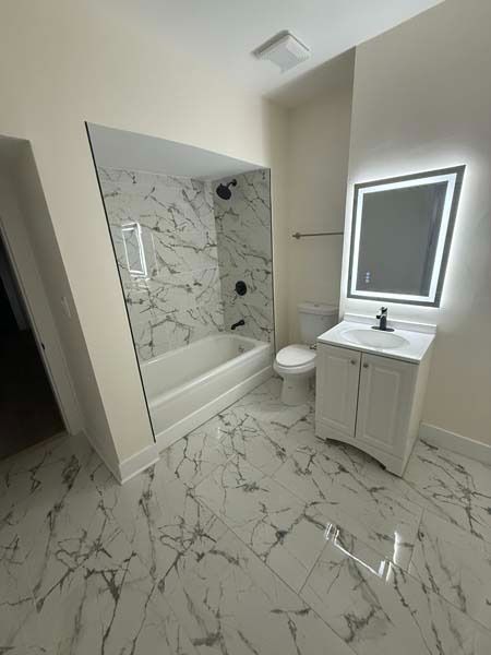 Modern bathroom with marble tile, bathtub, toilet, and lit vanity mirror