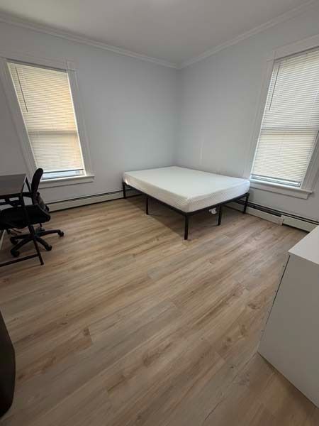 Empty bedroom with a bed, desk chair, and two windows with blinds and radiator heaters