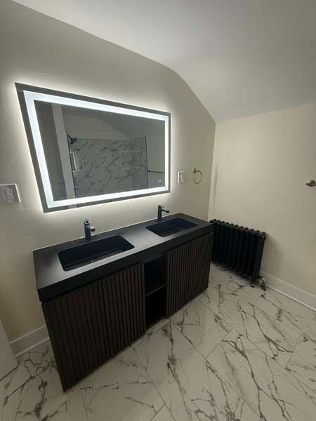 Modern bathroom vanity with dual sinks, lit mirror, white marble floor, and black ribbed cabinets
