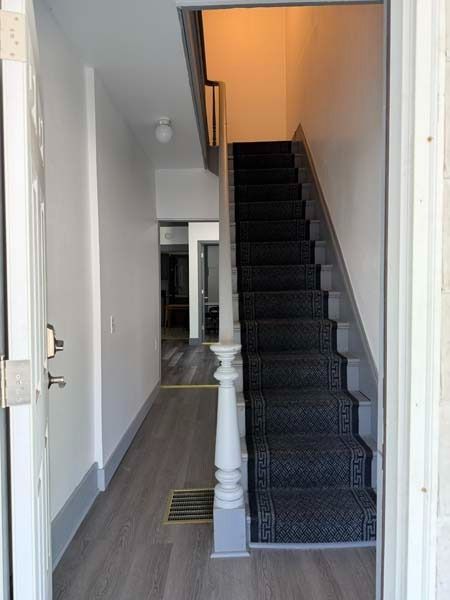 Narrow hallway with dark carpeted staircase, white walls, and a front door opening into the entryway