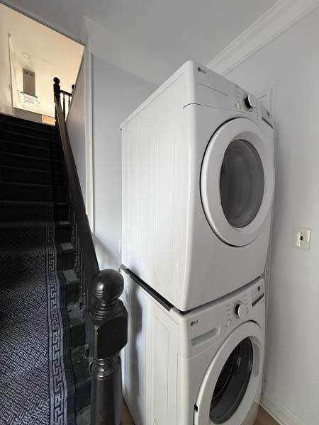 Stacked white washer and dryer in a narrow hallway beside a staircase.