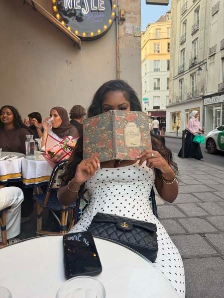 Woman in a white polka-dot dress holding a menu at an outdoor café table with a phone and black purse