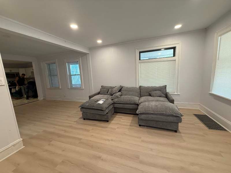 Modern living room with gray sectional sofa, light wood floors, recessed lighting, and windows with blinds