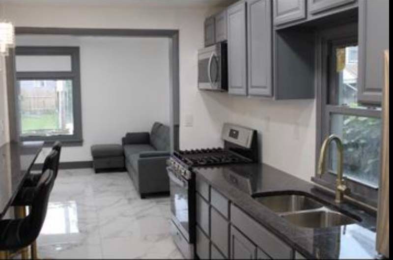 Modern kitchen with gray cabinets, black countertops, stainless sink, and a tiled floor leading to a living area.