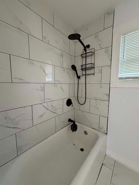 White-tiled bathroom with a bathtub, black shower fixtures, and a window with blinds