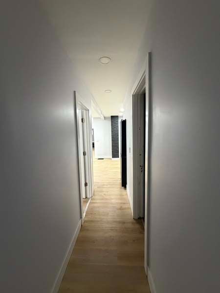 Narrow white hallway with light wood flooring and doors on both sides, leading to a bright room.