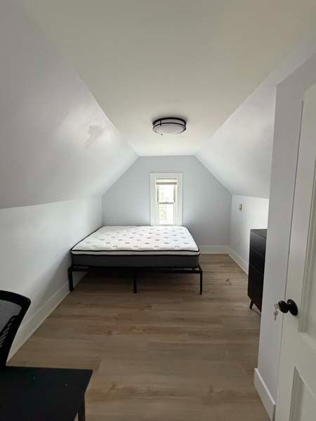 Narrow attic bedroom with a bed, white sloped walls, wood floor, and a small window at the far end