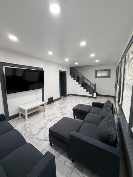 Bright modern living room with black sectional sofas, wall-mounted TV, white decor, and staircase in back