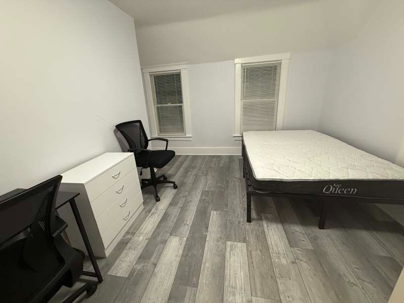 Small bedroom with a bed, desk, chair, dresser, and two windows on a gray wood floor.