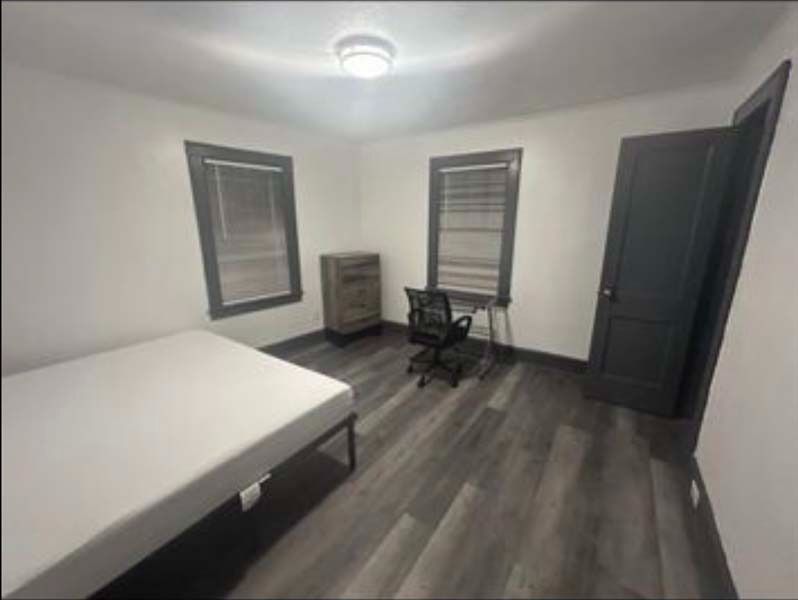 Empty bedroom with gray walls, dark flooring, bed, desk, chair, dresser, and two windows with blinds