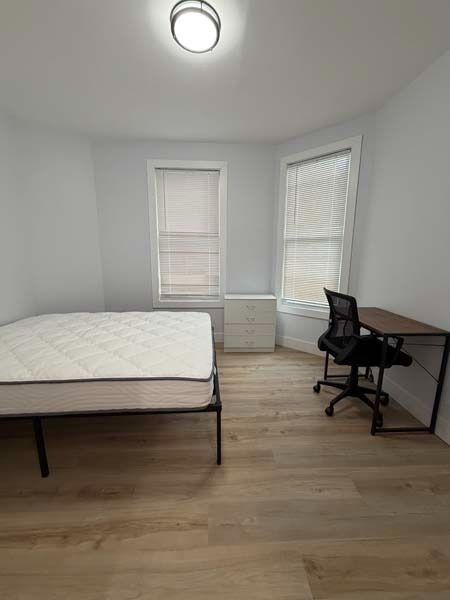 Sparse bedroom with bed, desk, chair, and two windows on light wood floors