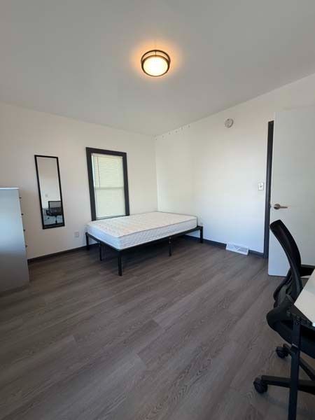 Empty bedroom with a bed, wall mirror, and office chair on a wooden floor.