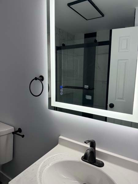 Modern bathroom vanity with lit mirror, black faucet, towel ring, and toilet visible in reflection