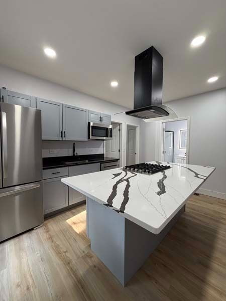 Modern kitchen with stainless steel appliances, white marble island, gray cabinets, and black range hood