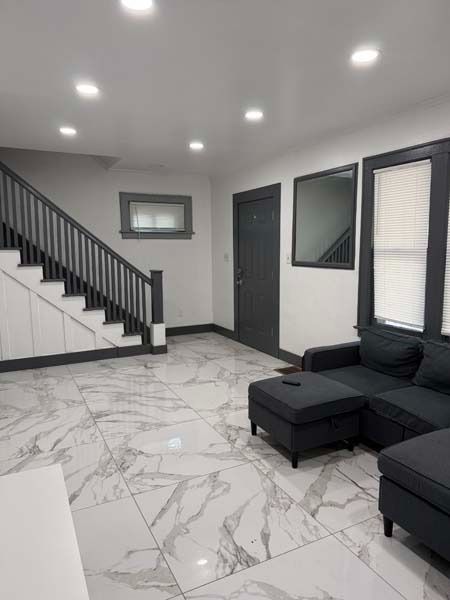 Modern living room with glossy white tile floor, black sectional sofa, staircase, and recessed ceiling lights