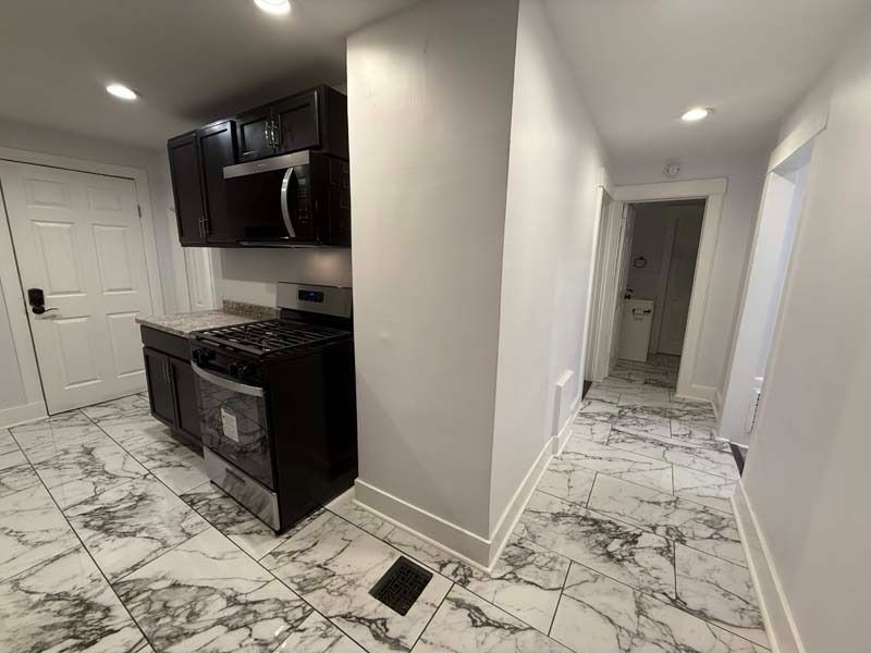 Empty modern kitchen with black appliances, white cabinets, and marble-patterned floor, view into hallway