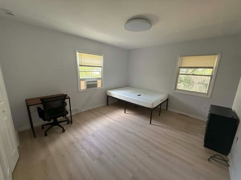 Empty bedroom with light wood floor, gray walls, two windows, bed, desk chair, and small cabinet.