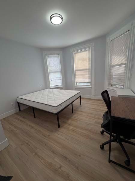 Empty bedroom with a bed, desk chair, and three windows in a bright white room.