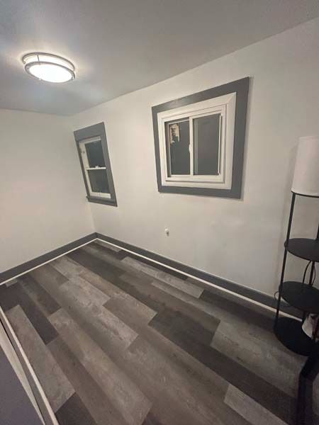 Small empty room with gray wood flooring, white walls, two windows, and a ceiling light.