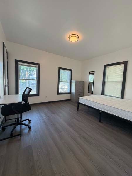 Empty bedroom with bed, desk chair, and three windows on dark wood floors