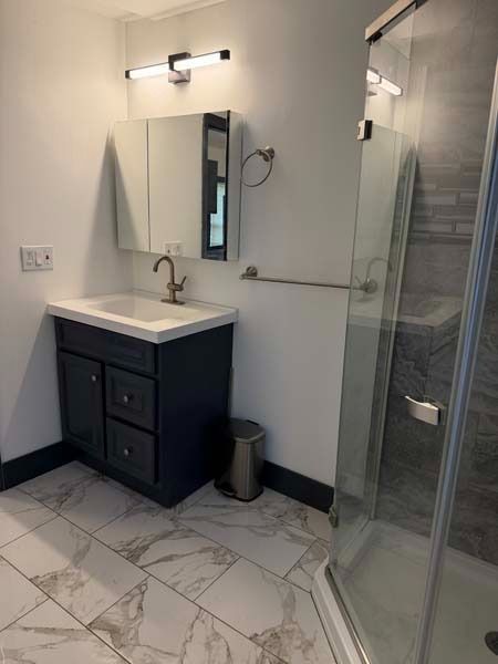 Small modern bathroom vanity with mirror, light, marble tile floor, and glass shower enclosure