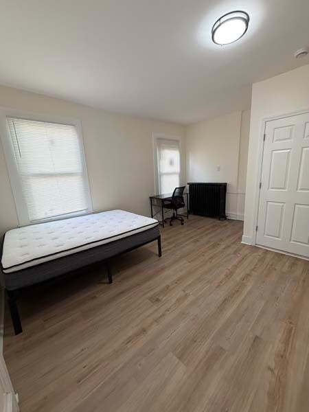 Sparse bedroom with a bed, desk, TV stand, closet doors, and two windows on wood floors