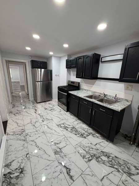 Modern kitchen with black cabinets, stainless steel fridge, white marble-look floor, and bright recessed lighting