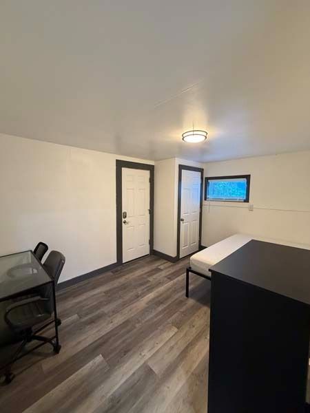 Small bedroom or office with a bed, desk, chair, two doors, and a small window.
