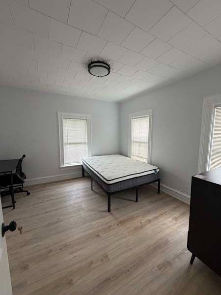 Empty bedroom with a twin bed, light wood floor, gray walls, and three windows.