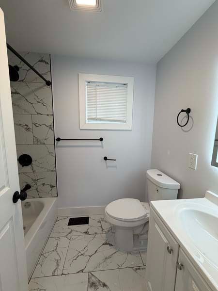 Small modern bathroom with white fixtures, marble tile, bathtub, toilet, and vanity.