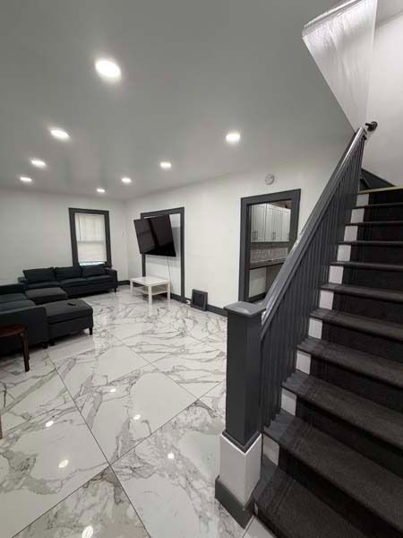 Modern living room with black sofas, wall-mounted TV, marble floor, and staircase with dark railing