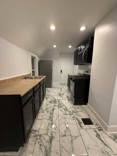 Empty kitchen with marble-pattern tile floor, black cabinets, white walls, and recessed ceiling lights.