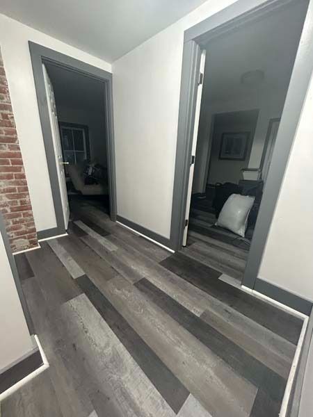 Hallway with gray wood-look flooring, white walls, open doorways, and a brick accent wall.
