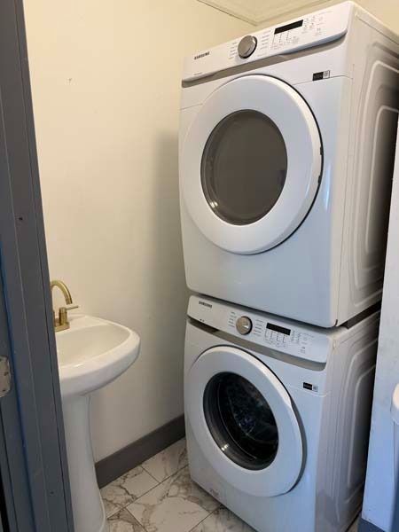 Stacked white washer and dryer in a narrow laundry room beside a small sink.