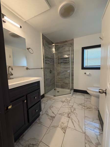 Modern bathroom with marble tile floor, glass corner shower, dark vanity, mirror, and a window.