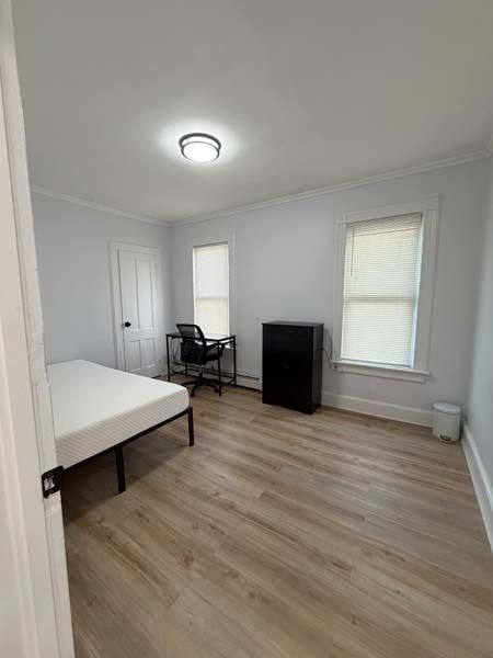 Small bright bedroom with bed, desk chair, dresser, and two windows on wood flooring.