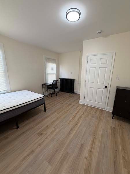 Empty bedroom with bed, desk, dresser, and white closet doors on light wood flooring.