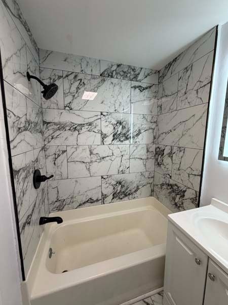 Small bathroom with marble shower walls, a white tub, and a sink beside a window.