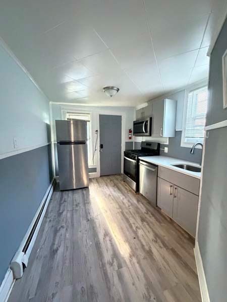 Narrow modern kitchen with stainless fridge, white cabinets, gray walls, and wood-look flooring.