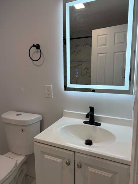 Small bathroom with white vanity, black faucet, lit mirror, toilet, and towel ring on the wall.