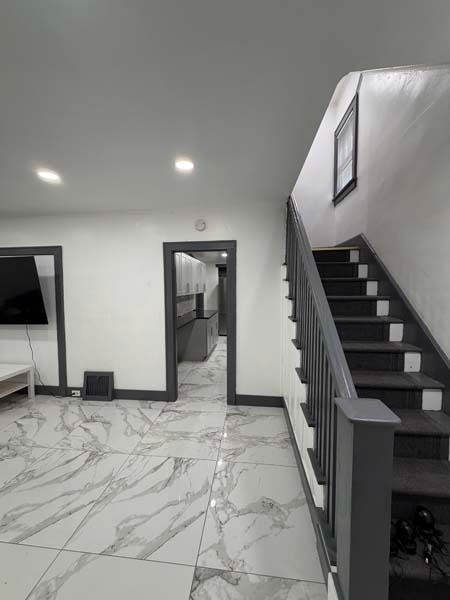 Modern living room with white marble floor, black staircase, wall-mounted TV, and doorway to kitchen.