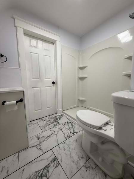 Bright white bathroom with toilet, tub, built-in shelves, and marble-pattern tile floor.