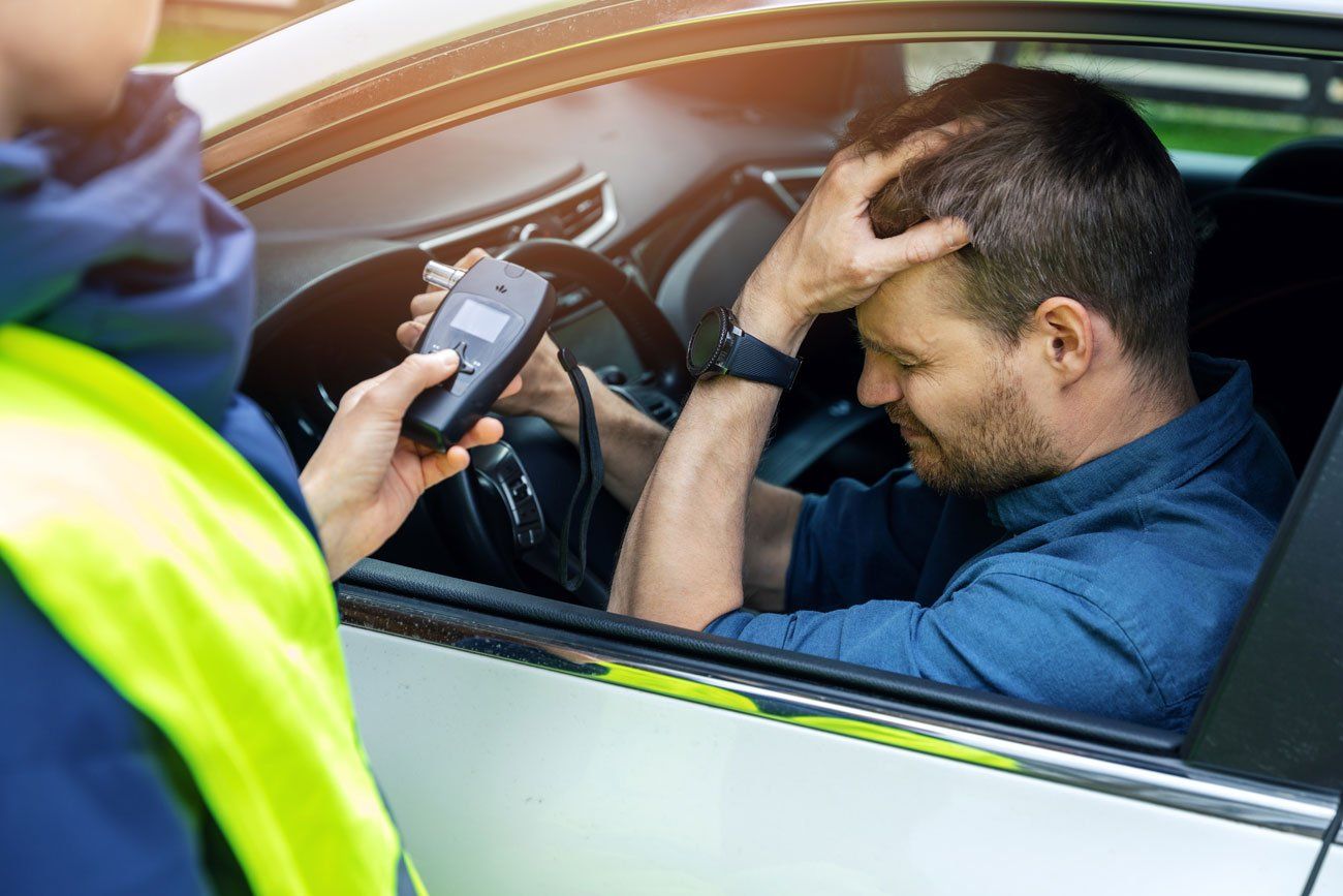 A police officer holds a breathalyzer for a stressed driver sitting in a car.