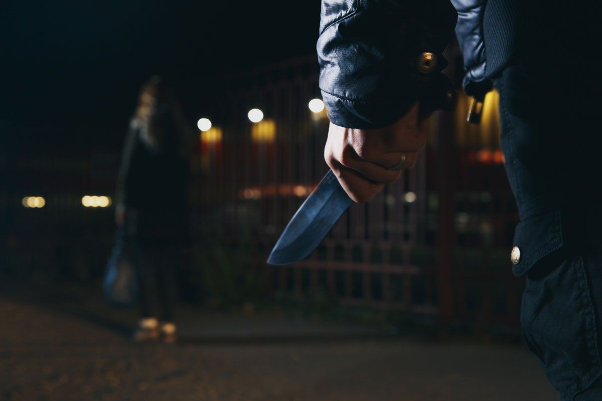 Close-up of a person holding a knife at night, with a blurred figure in the background.
