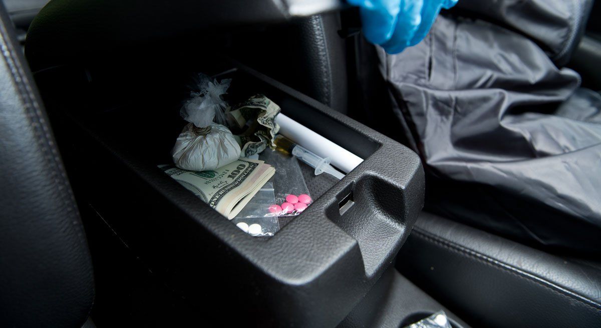 A gloved hand inspects a car’s center console containing a plastic bag of white powder, currency, and pills.