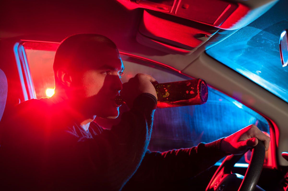 A person in a car drinks from a bottle, illuminated by flashing red and blue police emergency lights.
