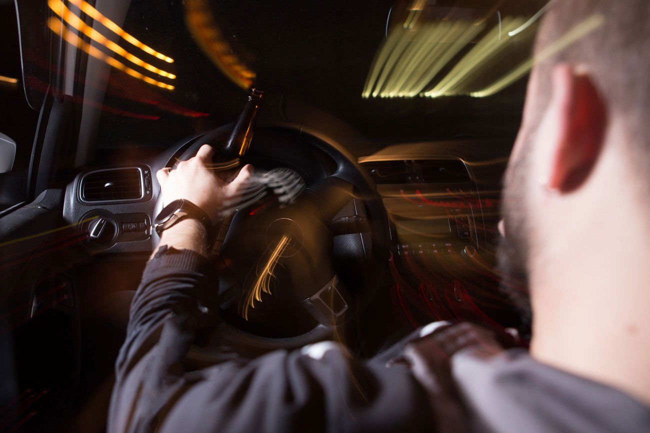 A person holds a bottle while driving a car at night, with motion-blurred street lights visible through the windshield.