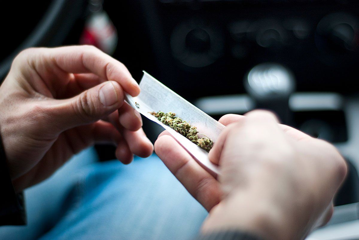 Close-up of hands preparing to roll a joint with dried cannabis inside a paper, with a car interior in the background.