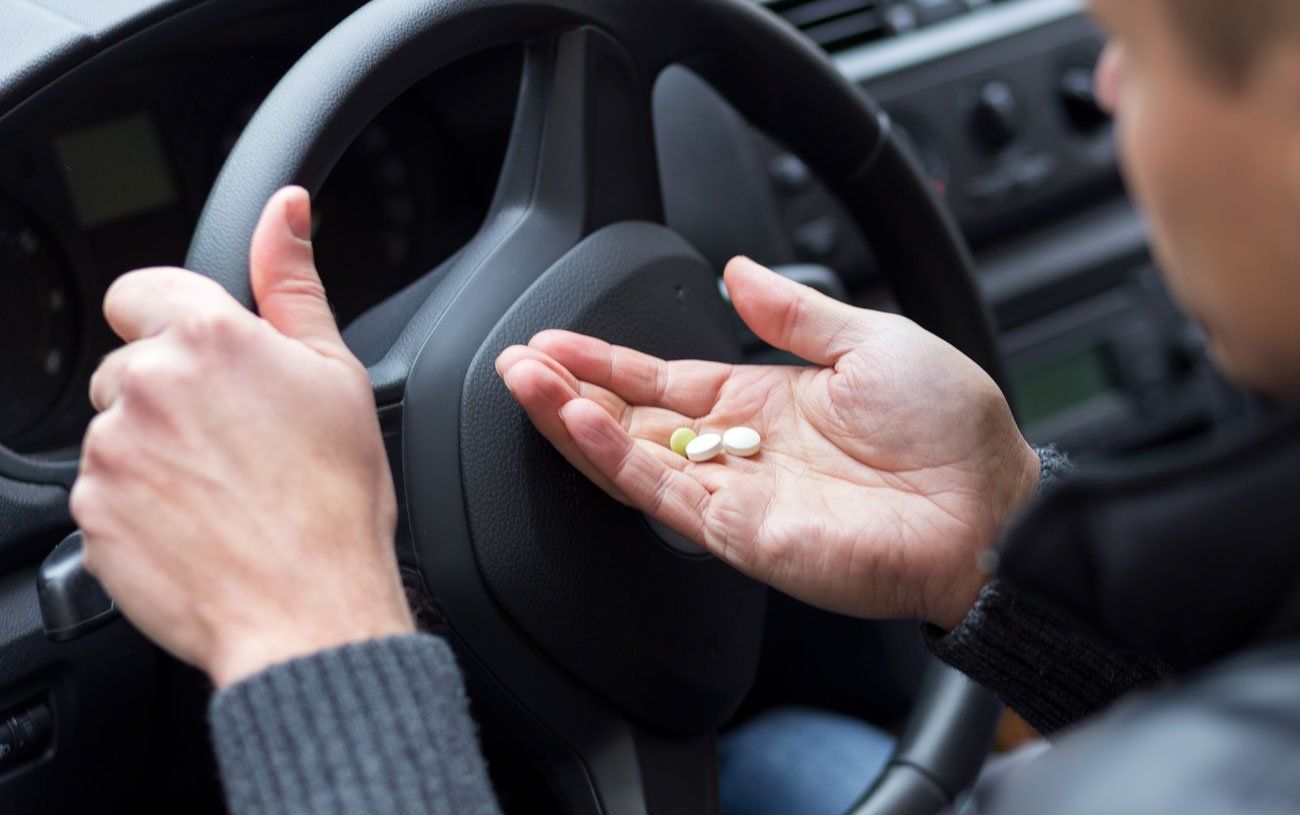 A person sitting in the driver's seat of a car holds several pills in the palm of their hand.