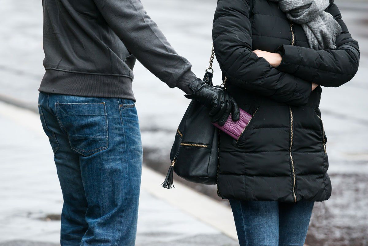 A person in a gray sweater and blue jeans uses a gloved hand to remove a purple wallet from another person's handbag.